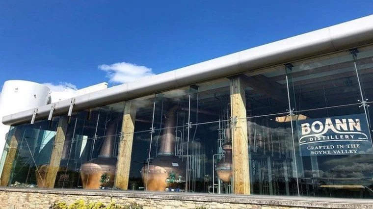 Copper pot stills visible through the glass facade of Boann Distillery under a blue sky
