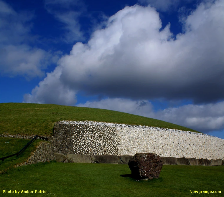 Newgrange passage tomb with white quartz facade, kerbstones and a standing stone on the lawn under a blue sky