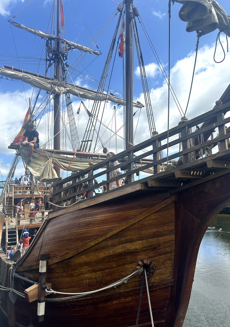 Bow of the Nao Santa María with bowsprit, furled sails and visitors on the foredeck against a blue sky